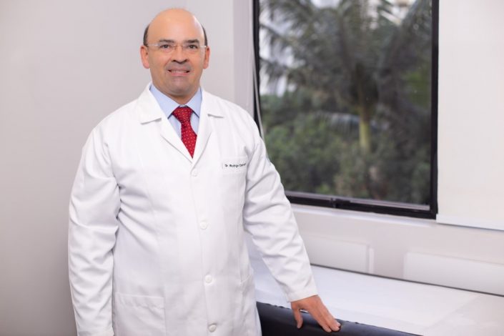 Retrato do doutor Rodrigo Campos, apoiando sua mão na maca dentro do consultório da Clínica Zequi