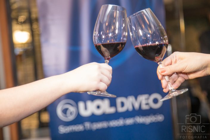 Nesta foto temos um brinde com taças de vinho em frente ao banner da Uol Diveo, cobertura fotográfica para evento corporativo em evento promovido no Le Bife